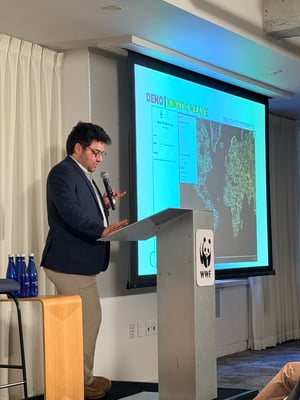 A presenter holds a microphone at a podium beside a projected map of the WWF Basin Threshold Tool during a live demonstration.